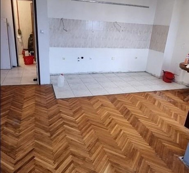 Room with herringbone parquet flooring leading to a tiled area intended as a space for kitchen installation, with wall-mounted electrical connections.